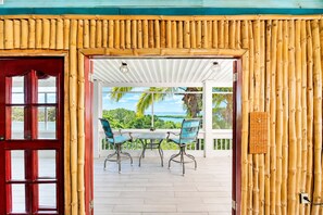 A covered outdoor seating area with a table and chairs, a view of a tropical landscape, and a bamboo wall.