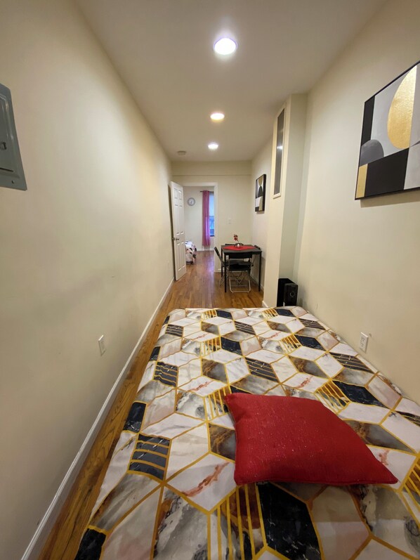 A narrow hallway with a bed at the end, a door leading to another room, and a small table with a chair.