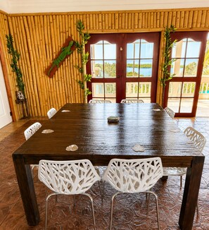 A dining area with a long wooden table, white chairs, and a view through large windows. - Vistas 360 Yellow - 3 Bedroom Home W/ Ocean Views (Politilly Bight)