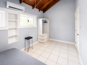 A bedroom with a bed, a white wardrobe, a small black table, a white air conditioning unit, and a window with a view of greenery. - Vistas 360 Yellow - 3 Bedroom Home W/ Ocean Views (Politilly Bight)