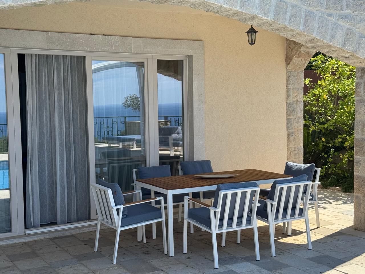 Un patio exterior con mesa de madera y sillas blancas, una puerta corrediza de vidrio y un muro de piedra.