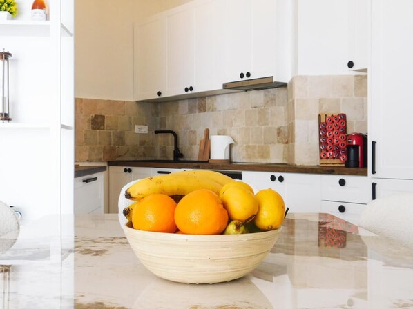 A kitchen with a marble countertop, a bowl of fruit, and white cabinets.