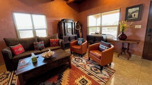 A cozy living room with a brown leather sofa, two armchairs, a wooden coffee table, and a patterned rug. There are two windows with blinds, a wooden cabinet, and a small table with a vase of flowers.