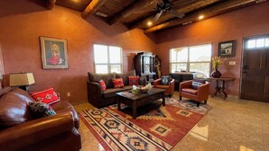 A spacious living room with a leather sofa, a coffee table, and a television. There are two windows providing natural light, and a wooden ceiling with a fan.