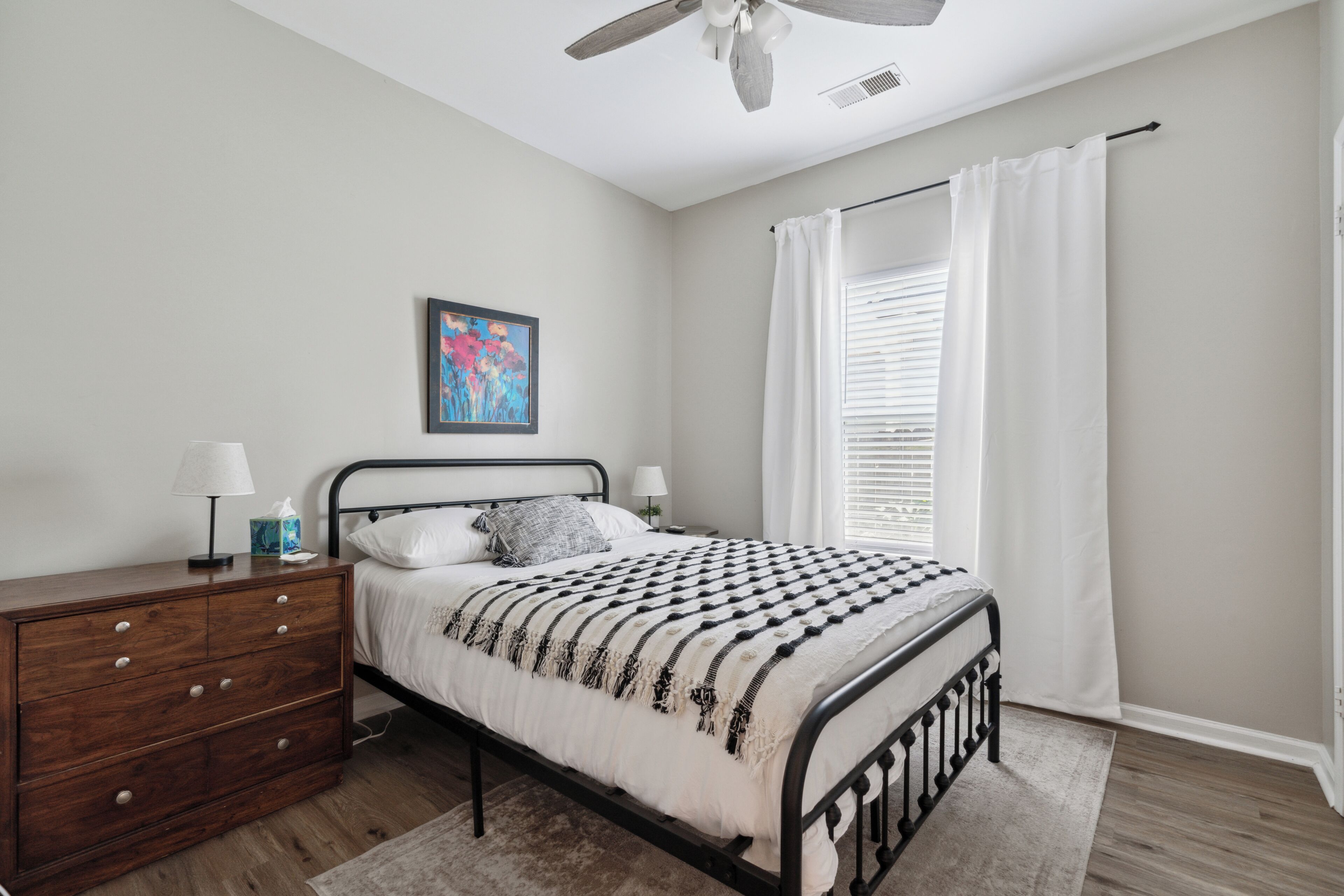 A bedroom with a bed, wooden dresser, bedside lamps, a ceiling fan, and a window with white curtains.