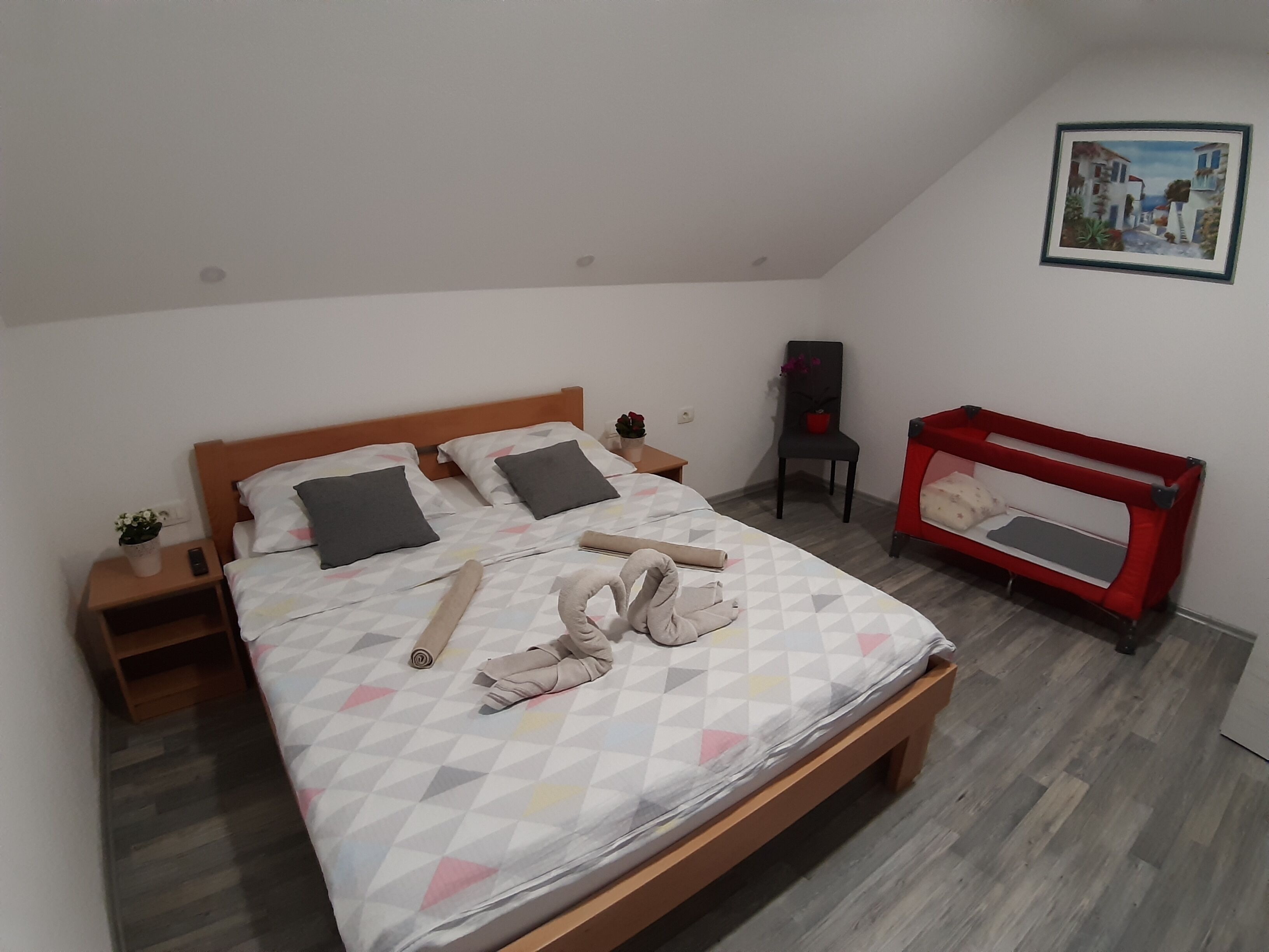 Una cama bien hecha con almohadas, una cuna roja para bebé, una silla y una mesita con una planta y un control remoto.