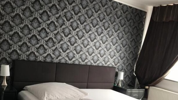 A neatly made bed with white linens and a folded towel, a dark headboard, and a patterned wallpaper. - Pension Alis (Augsburg)