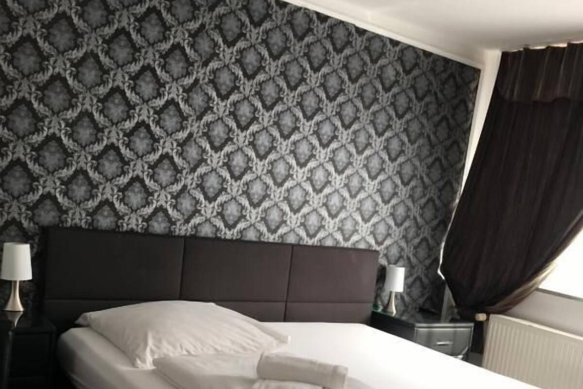 Ein ordentlich bezogenes Bett mit weißen Leinen und einem gefalteten Handtuch, ein dunkles Kopfteil und eine Wand mit Muster.