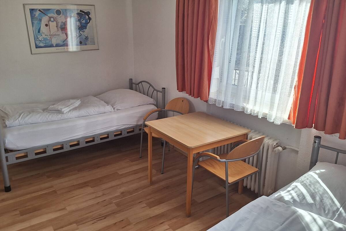 Ein kleines Zimmer mit einem Einzelbett, einem Holztisch und Stuhl, einem Heizkörper und einem Fenster mit roten Vorhängen.