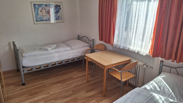A small, single-bed room with a wooden table and chair, a radiator, and a window with red curtains.