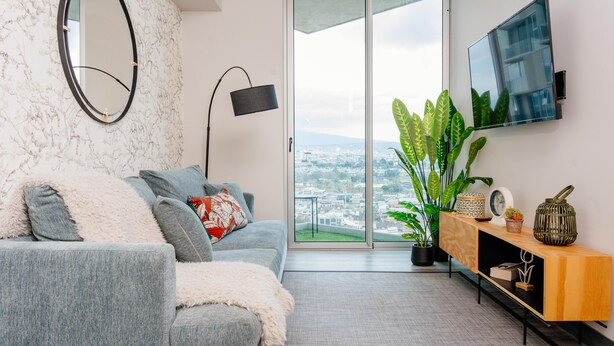 A modern living room with a grey sofa, a black floor lamp, a wooden console table, and a large window offering a city view. - Secrt Sabana 2BR · City View · High Floor · Pool & Gym · Apt 2416 (Mata Redonda)