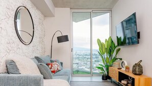 A modern living room with a grey sofa, a round mirror, a floor lamp, and a TV on the wall. - Secrt Sabana 1BR · Balcony Workspace · City View · Parking · Apt 2915 (Mata Redonda)