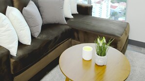 A modern living room with a dark brown leather sofa, white and gray cushions, a small round wooden coffee table with a white vase and a candle, and a large window offering a city view. - Secrt Sabana 2BR · Skyline View · High Floor · Pool & Gym · Apt 2810 (Mata Redonda)