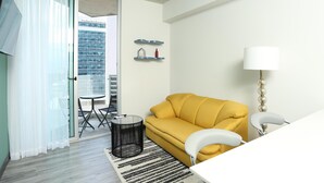 Un salon moderne avec un canapé jaune, un fauteuil blanc, une petite table ronde et une lampe. Une porte vitrée donne accès à un balcon d’où l’on peut admirer un immeuble.