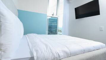 Cama bem arrumada com roupa de cama branca em quarto com parede azul e televisão fixada na parede.