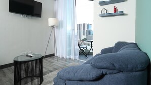 Una sala de estar moderna con un televisor de pantalla plana, una mesa redonda de vidrio, una lámpara de pie, un sofá y un balcón con vistas a edificios.