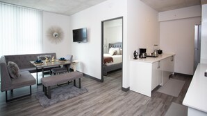 A modern hotel room with a grey sofa, a dining table, a small kitchen area, and a large mirror.