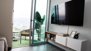 A modern living room with a large TV, a sliding glass door leading to a balcony with a view of the city, a small table, and a chair. - Secrt Sabana 1BR · High Floor · Workspace + WiFi · Parking · Apt 2006 (Mata Redonda)
