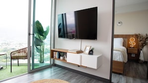 A modern living room with a large TV, a sliding glass door leading to a balcony, a wooden console table, and a bed with a nightstand. - Secrt Sabana 1BR · High Floor · Workspace + WiFi · Parking · Apt 2006 (Mata Redonda)