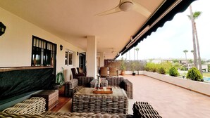 Un área de estar techada al aire libre con mobiliario de mimbre, ventilador de techo y vistas a palmeras y una piscina.