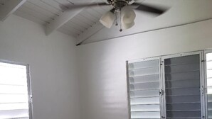 Un dormitori amb llit, ventilador de sostre i dues finestres amb persianes.