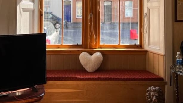 Una habitació amb un gran finestral, un coixí amb forma de cor sobre una banca i una televisió sobre un suport de fusta.