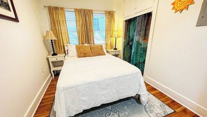 A neatly arranged bedroom with a bed, two bedside tables, a nightstand, a window with curtains, and a wall-mounted sun decoration. - Central Location  Beach Access BBQ  Patio Cottage (Long Beach)