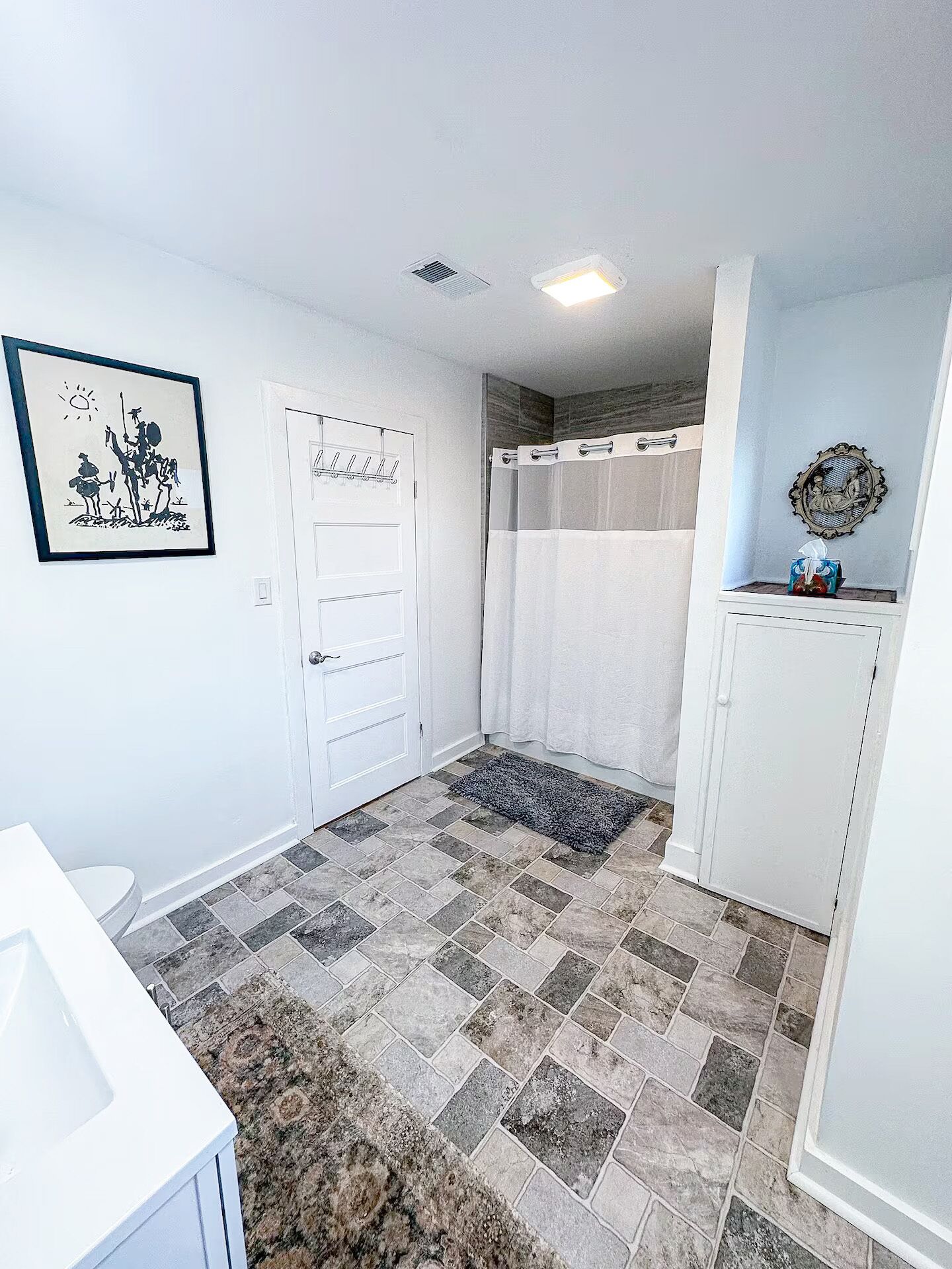 Un baño con una puerta blanca, cortina de ducha, una alfombra pequeña y un cuadro en la pared.