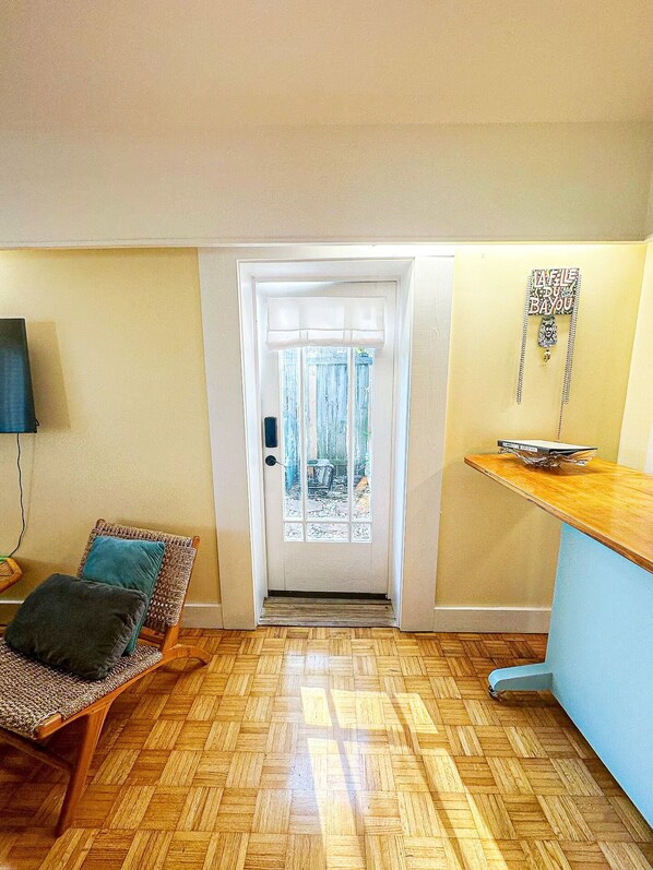 Una habitación con piso de madera en patrón espiga, un sillón de mimbre con cojines, una mesa pequeña con un objeto decorativo y una puerta que da a un patio trasero.