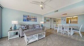 A living room with a sofa, coffee table, and a dining area with chairs and a table. There is a ceiling fan, a painting on the wall, and a kitchen area visible through an open door. - Anchorage 206 (Sarasota)