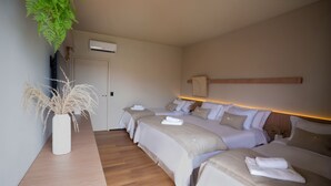 Habitación de hotel con tres camas, una mesita de noche de madera, un jarrón blanco con plantas secas y un equipo de aire acondicionado en la pared.