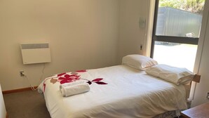 A single bed with a floral bedspread, a white pillow, and a rolled-up towel. There is a power outlet on the wall and a window with a view of the outside.