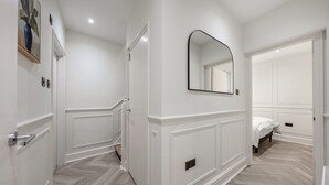 A modern, well-lit hallway with white walls and a herringbone-patterned floor. There is a door on the left, a mirror on the right, and a bed in the background. - Pimlico Brand New Luxury Home (London)