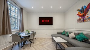 A modern living room with a dining area, a grey sofa, a flat-screen TV displaying 'NETFLIX', and a colorful abstract painting on the wall. - Pimlico Brand New Luxury Home (London)