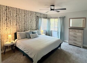A bedroom with a bed, a bedside table with a lamp, a dresser, a ceiling fan, and a window with curtains.