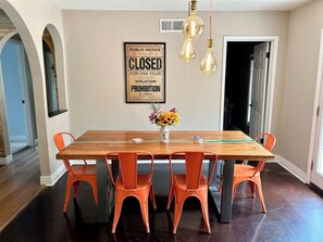 Un comedor con una mesa de madera y sillas anaranjadas, un aviso enmarcado en la pared y un jarrón con flores.