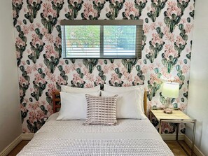 Un dormitorio con cama, almohadas, una mesita de noche con lámpara y una ventana con persianas.