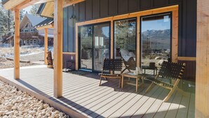 Une terrasse en bois avec deux chaises et une table en bois, une cheminée en pierre et une grande porte vitrée donnant sur un espace abrité.