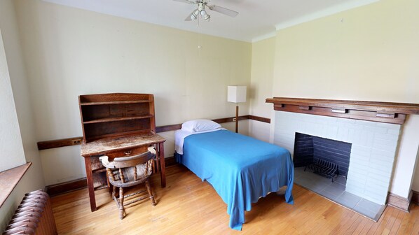 A room with a wooden desk, chair, and a bed with a blue bedspread. There is a white wall and a fireplace with a white brick surround.