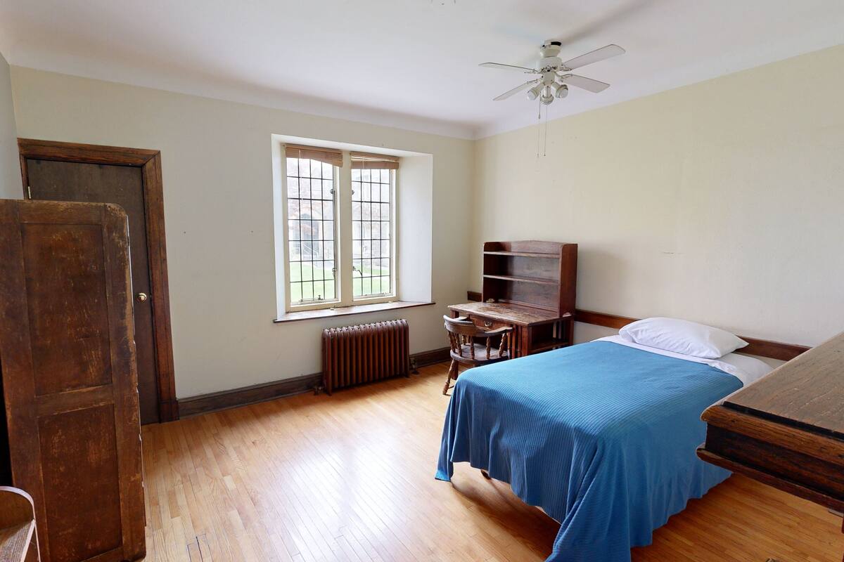 A room with a bed, a wooden desk, a radiator, a window, and a ceiling fan.