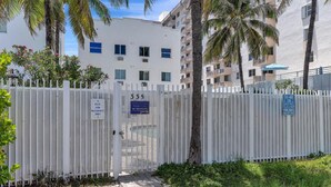 A gated residential area with a white fence, palm trees, and a building with multiple balconies. - BeachFront On The Sand Unit #326 South of 5th, 335 Ocean Dr, Miami Beach (Miami Beach)