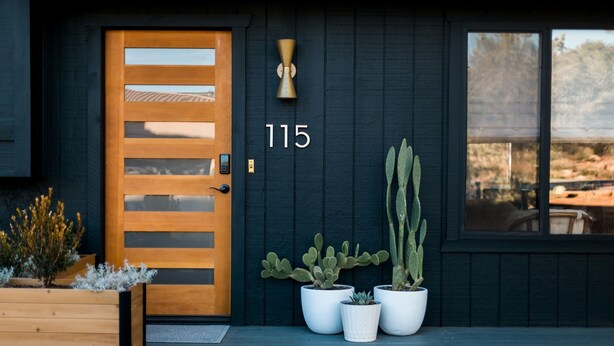 Une maison noire avec une porte en bois, le numéro 115, et trois cactus en pot sur le porche.