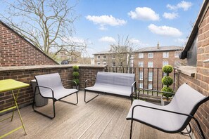 A rooftop terrace with a brick wall, two grey chairs, a yellow table, and a view of buildings in the background.
