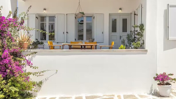 A covered outdoor seating area with a wooden table and chairs, surrounded by potted plants and a white wall. - Naousa | 2BR House in the Heart of Town | Citrine (Naousa)