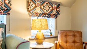 A room with a window, a table with a lamp, a brown leather chair, and a green armchair.