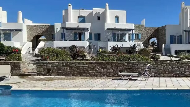 A poolside view of a resort with white buildings, stone walls, and a clear blue sky. - Ampelas | 3BR Cycladic House | White Heaven Apt4 (Ampelas)