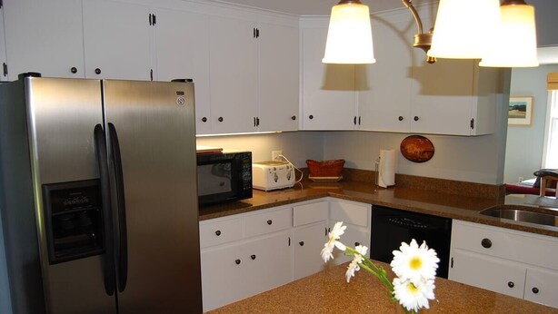 A kitchen with white cabinets, a stainless steel refrigerator, a microwave, a toaster, and a vase with flowers on the counter. - Captain's Quarters Cottage at Dockside Guest Quarters in York Harbor,  Maine. (YORK)