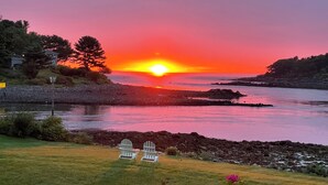 A sunset over a coastal area with a grassy foreground and two Adirondack chairs. - Captain's Quarters Cottage at Dockside Guest Quarters in York Harbor,  Maine. (YORK)