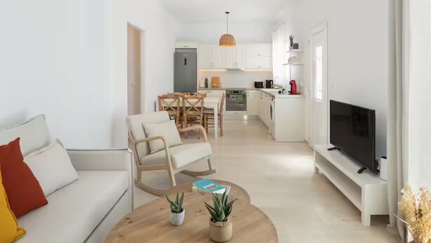 A modern living room with a sofa, coffee table, and TV. There is a kitchen in the background with white cabinets and a dining area. - Naousa | 2BR Cycladic Town-House | Jasper (Naousa)