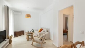 A modern living room with a sofa, a wooden rocking chair, a coffee table, and a TV. - Naousa | 2BR Cycladic Town-House | Jasper (Naousa)